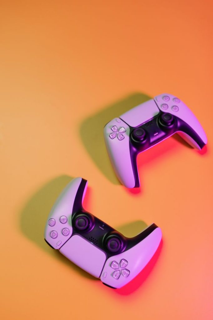Two white video game controllers on an orange background, shot from above with colored lighting.